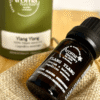 Brown glass bottle of Ylang Ylang essential oil with a dropper cap, placed on a woven mat, with a green Aroma bottle in the background.